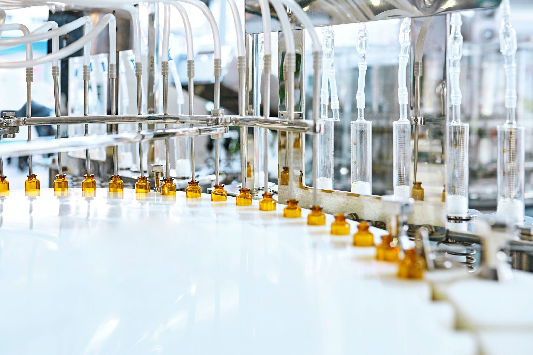 Landscape oriented 5700 pixel wide by 3800 pixel high jpg photo of a pharmaceutical assembly line where medicine is squeezed through plastic tubes into amber-colored glass vials.